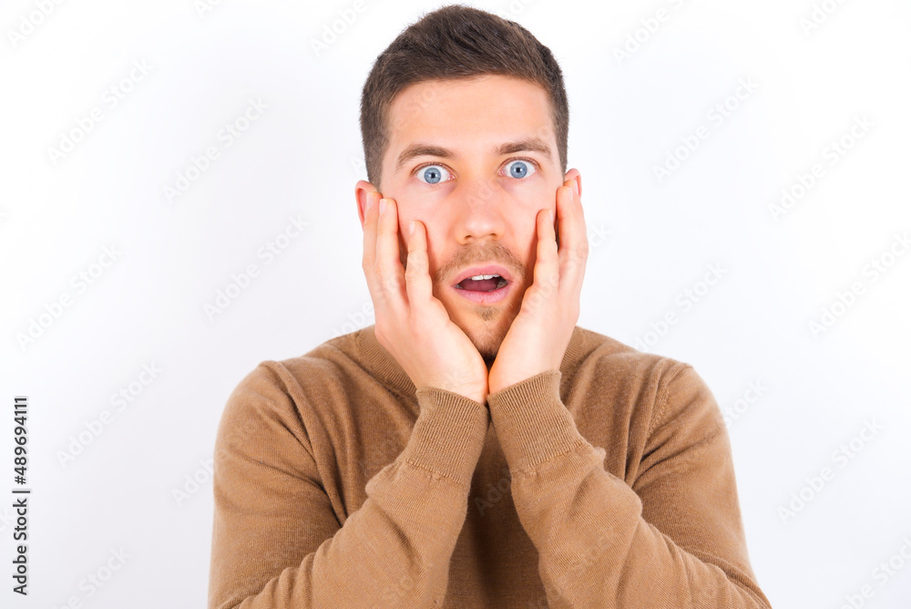 Stupefied young caucasian man wearing grey turtleneck over white background expresses excitement and thrill, keeps jaw dropped, hands on cheeks, has eyes popped out Stupefied young caucasian man wearing grey turtleneck over white background expresses excitement and thrill, keeps jaw dropped, hands on cheeks, has eyes popped out