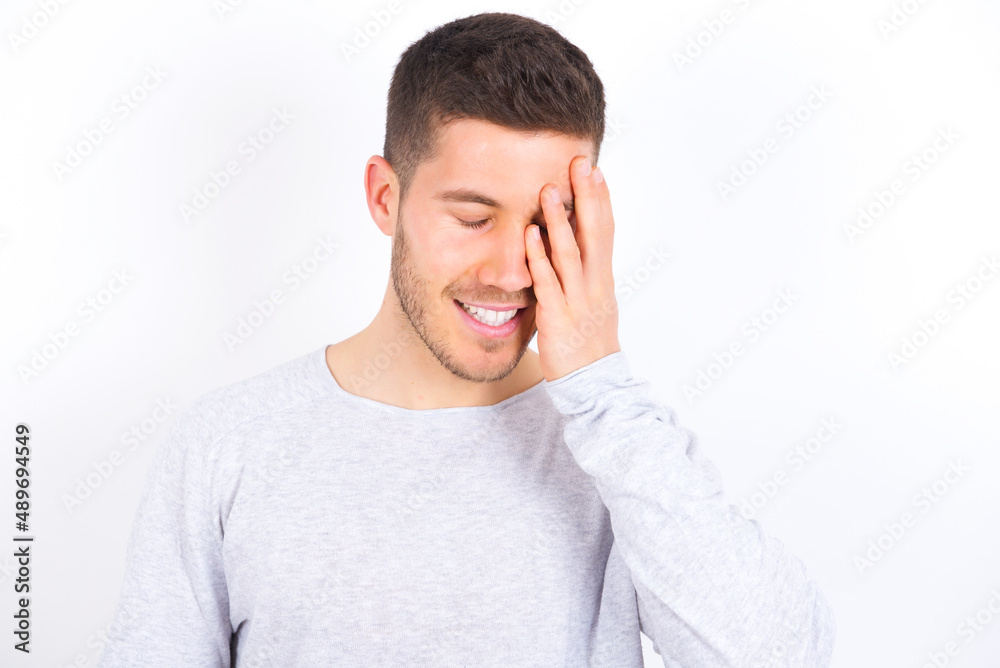 young caucasian man wearing grey sweater over white background makes face palm and smiles broadly, giggles positively hears funny joke poses young caucasian man wearing grey sweater over white background makes face palm and smiles broadly, giggles positively hears funny joke poses