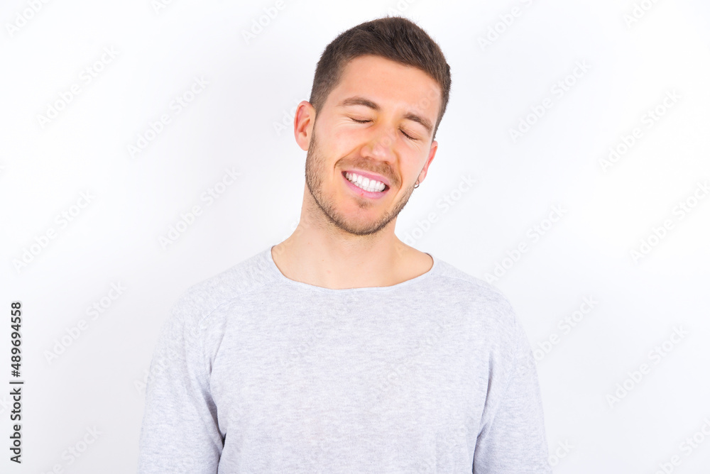 Fototapeta premium Positive young caucasian man wearing grey sweater over white background with overjoyed expression closes eyes and laughs shows white perfect teeth