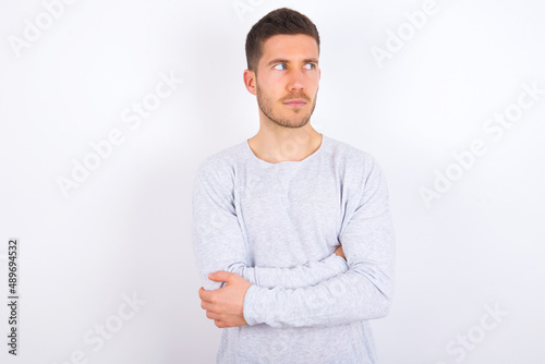 Pleased young caucasian man wearing grey sweater over white background keeps hands crossed over chest looks happily aside