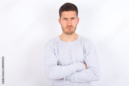 Gloomy dissatisfied young caucasian man wearing grey sweater over white background looks with miserable expression at camera from under forehead, makes unhappy grimace