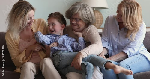 Multigenerational family play sit on sofa at home. Elderly and young relatives female enjoy carefree weekend together. Young mom, grandmother, great-grandma tickling little girl. Bonding, fun concept