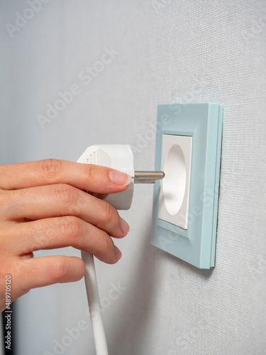 Close-up of a woman's hand holding a white plug and about to plug it into a blue outlet on the wall in the house. The concept of energy conservation. Side view, selective focus