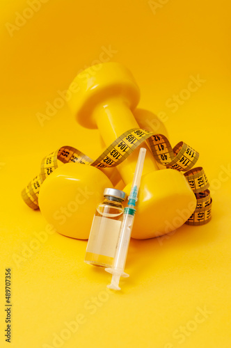 syringe and a jar of liquid lie on dumbbells with a measuring tape on a yellow background, vertical photo. the concept of doping in sports, steroids, testosterone and other drugs for weight loss.