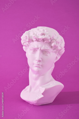 copy of the head of an antique statue of David in pink neon light on a purple background, trend colors © Natasha