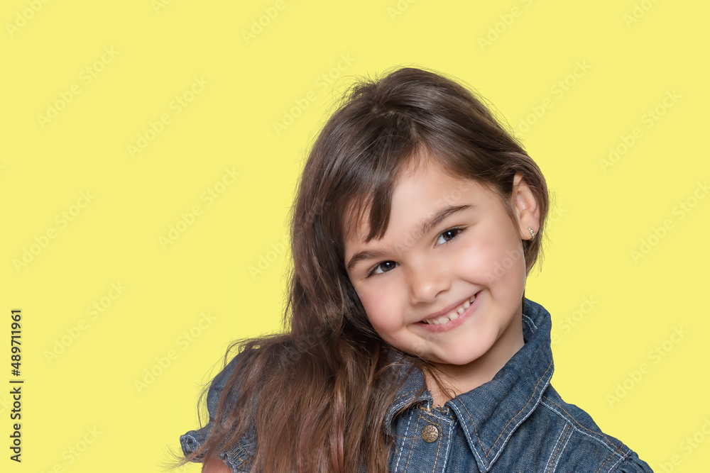 Smiling tanned long haired little girl wears denim costume is posing on yellow background. Smiling tanned long haired little girl wears denim costume is posing on yellow background.