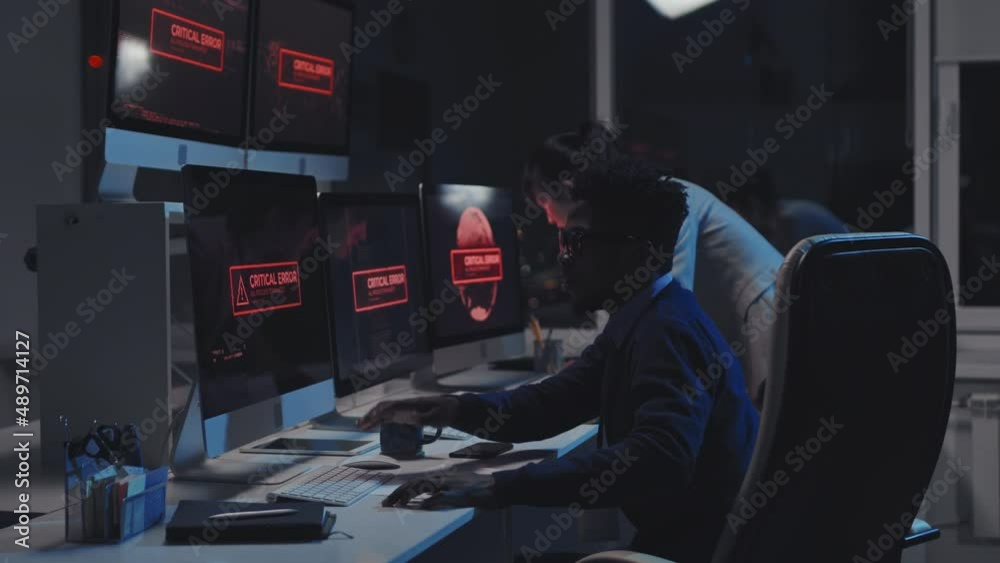 Side-view of two young diverse cyber security specialists fixing critical error, warning alarm showing on multiple computers monitors on desk in office at night