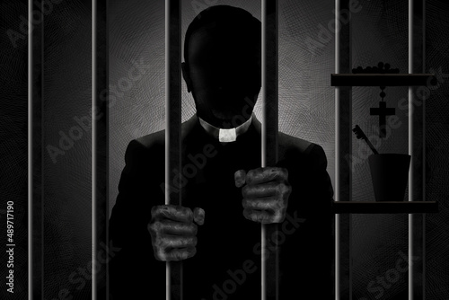 A Catholic priest identified by his clergyman collar is seen in silhouette behind bars with his filthy hands gripping the bars. He is in shadows and his face is in darkness. photo