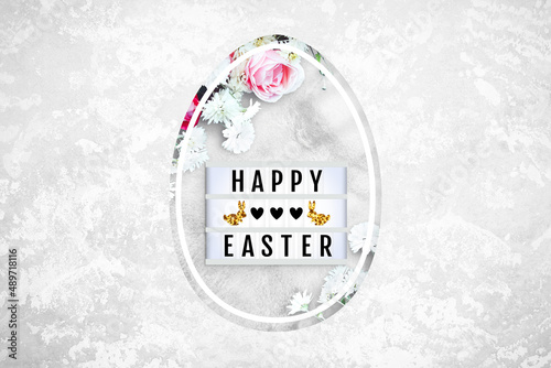 Lightbox and flowers in Easter egg shaped hole on a concrete background. Red and pink roses, white daisies. Lightbox with the lettering Happy Easter, hearts, bunnies. Top view, close up, copy space