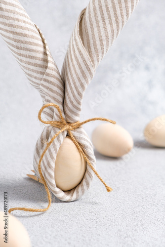 Close-up of Easter bunny made from egg and linen striped napkin. Easter eggs are light beige on light gray background. Trend of decorating traditional spring holiday Easter. Vertical, pastel colors.