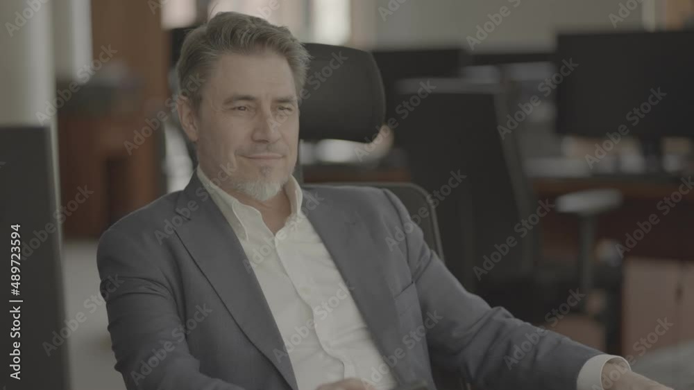 Business portrait - confident businessman sitting at desk in office. Happy mid adult man in shirt and jacket, smiling. Bearded, gray hair. Video in LOG format - S-LOG3 Cine.
