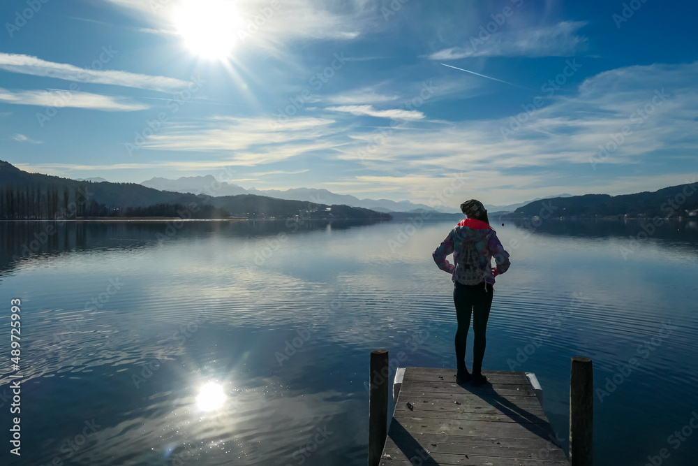 Happy woman standing on the pier at beautiful Woerthersee in Poertschach, Carinthia, Austria. Scenic lake landscape surrounded by Karawanks Alps. Fresh and clean air. Reflection in Lake Woerth