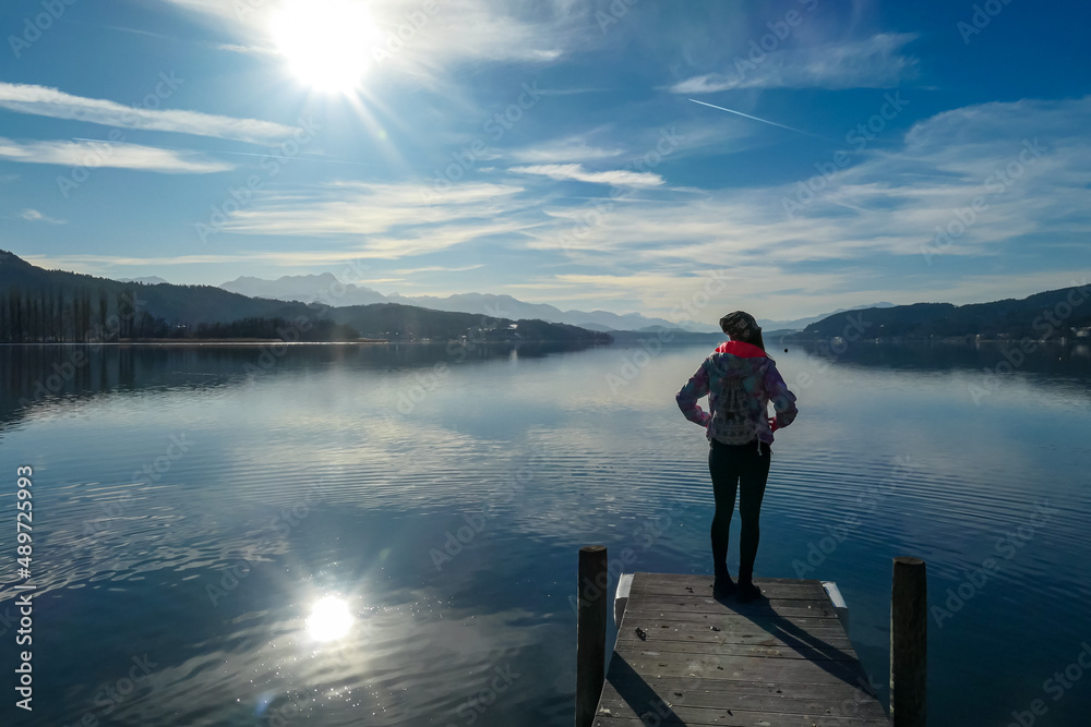 Happy woman standing on the pier at beautiful Woerthersee in Poertschach, Carinthia, Austria. Scenic lake landscape surrounded by Karawanks Alps. Fresh and clean air. Reflection in Lake Woerth Happy woman standing on the pier at beautiful Woerthersee in Poertschach, Carinthia, Austria. Scenic lake landscape surrounded by Karawanks Alps. Fresh and clean air. Reflection in Lake Woerth