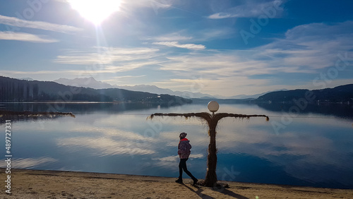 Happy woman walking along the shore of beautiful Woerthersee in Poertschach, Carinthia, Austria. Scenic lake landscape surrounded by Alps. Serenity. Fresh and clean air. Reflection in Lake Woerth