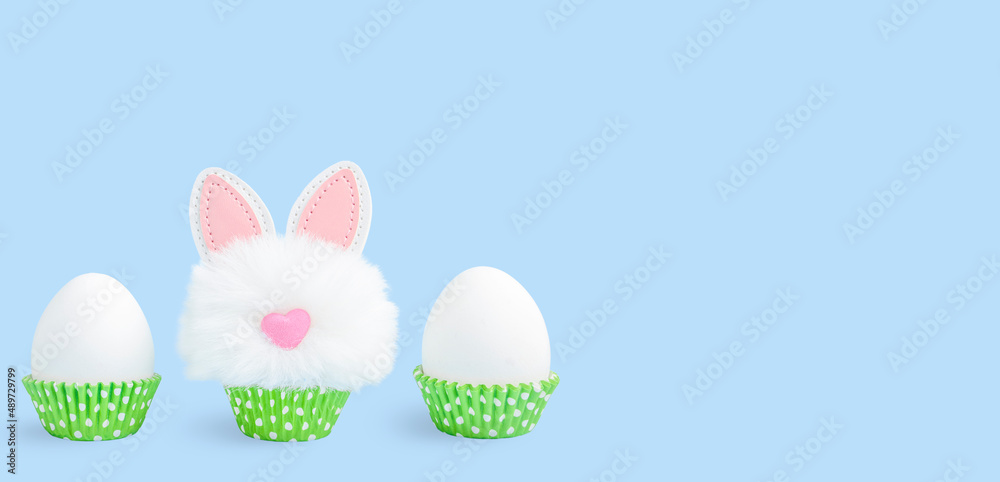 Unpainted Easter eggs in green paper cake pans and funny easter bunny toy on blue background. Banner with copy space Unpainted Easter eggs in green paper cake pans and funny easter bunny toy on blue background. Banner with copy space