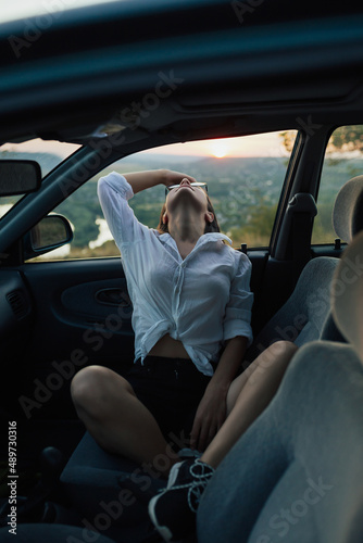 Young stylish beautiful woman sitting in her car in rural road. Perfect trip in summer time.