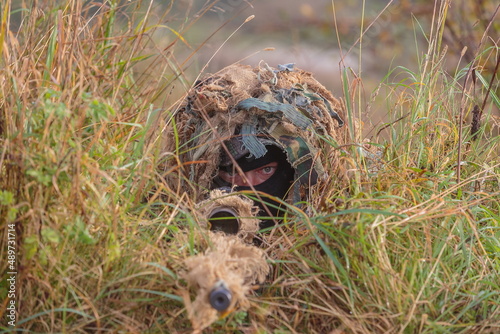 Sniper with Ghillies camouflage lying behind a sniper rifle with optics in the grass, illustration photo for war