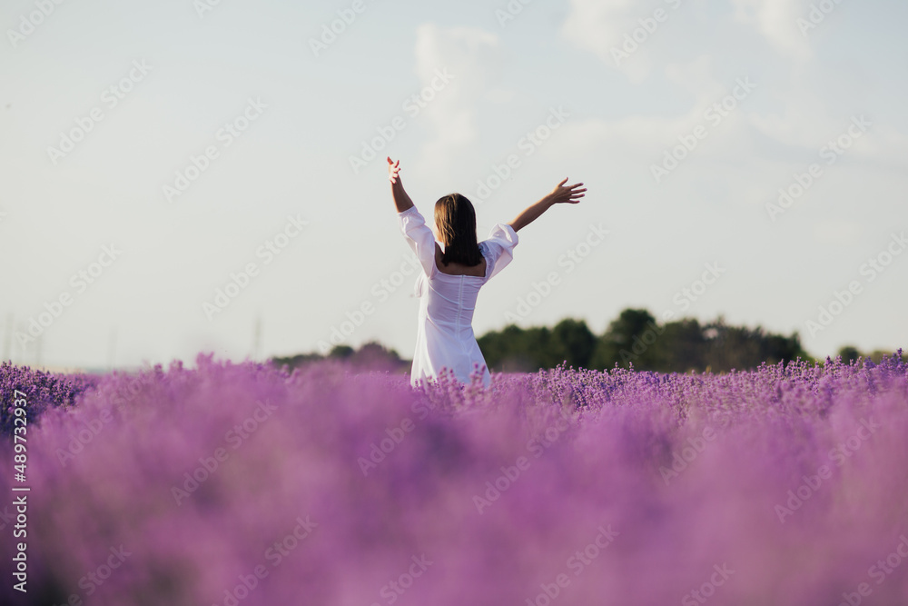 Beautiful young woman with a white dress standing in the middle of a lavender field with hands up.