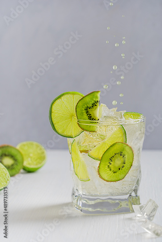 Homemade cold refreshing lemonade with ice cubes garnished with slices of fresh lime and ripe kiwi fruit with falling or levitating drops of drink served in glass on white wooden table at summer
