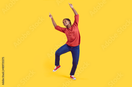 Happy cheerful young black girl having fun in the studio. Funny African American woman in comfortable casual clothes dancing against a vivid, vibrant yellow colour background in a modern photo studio © Studio Romantic