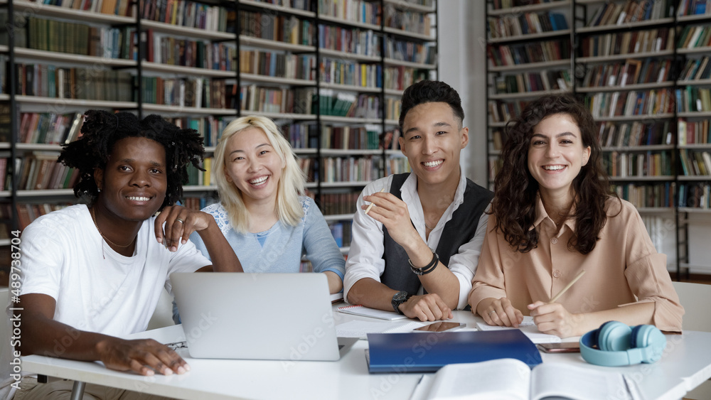 Portrait of smiling multiracial young students sitting at table with books and laptop, ready for online lesson in library or modern classroom. Joyful diverse classmates working together on project. Portrait of smiling multiracial young students sitting at table with books and laptop, ready for online lesson in library or modern classroom. Joyful diverse classmates working together on project.