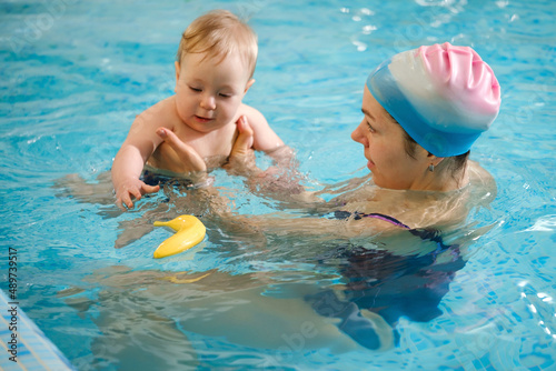 Early age swimming in pool. Baby boy trained to swim in water. Happy child with trainer woman in indoor swimming pool playing and having fun. Healthy and sport family with infant, active parent