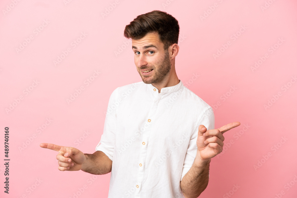 Young caucasian handsome man isolated on pink background pointing finger to the laterals and happy