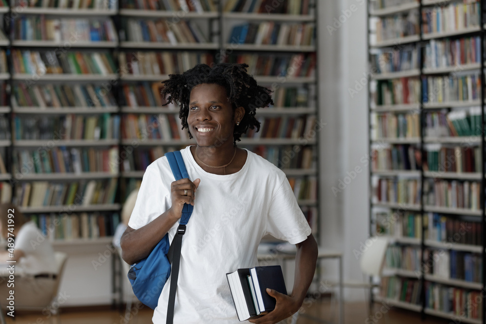 Happy motivated millennial African American student guy with dreads and backpack holding books in hands, looking in distance standing in college library, thinking of future career or university exams. Happy motivated millennial African American student guy with dreads and backpack holding books in hands, looking in distance standing in college library, thinking of future career or university exams.