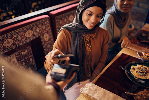 Happy Muslim woman paying contactless with mobile phone in restaurant.