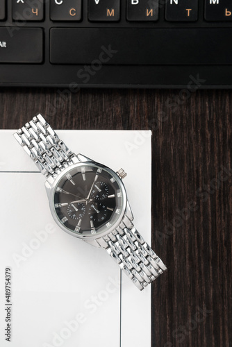 Wristwatch with a silver bracelet and a black dial lies on a book next to the keyboard on a dark wood table