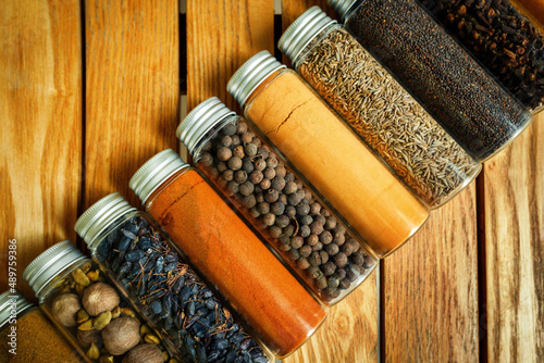 Different multi-colored spices in transparent jars lie on a wooden background.