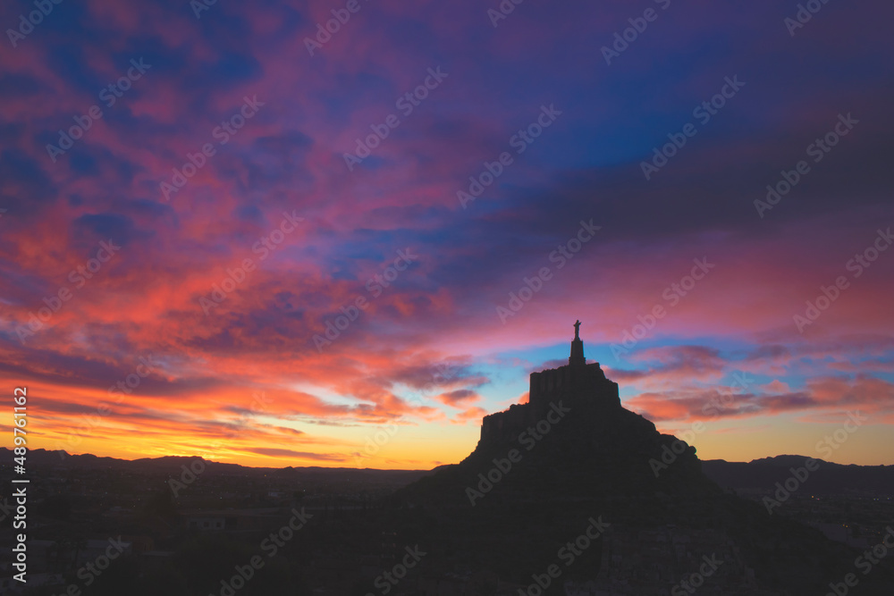 Silhouette of the medieval castle of Monteagudo, Murcia, Spain, with a cross on top. The sky shows the colors of dawn coloring the clouds above the sky