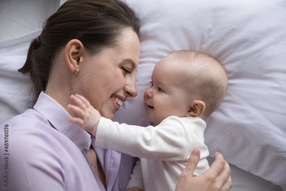 Happy young mother and cute cheerful baby relaxing in bedroom together. New mom lying on bed with pale linen, cuddling, hugging adorable infant, kid touching mum face. Top view, close up Happy young mother and cute cheerful baby relaxing in bedroom together. New mom lying on bed with pale linen, cuddling, hugging adorable infant, kid touching mum face. Top view, close up
