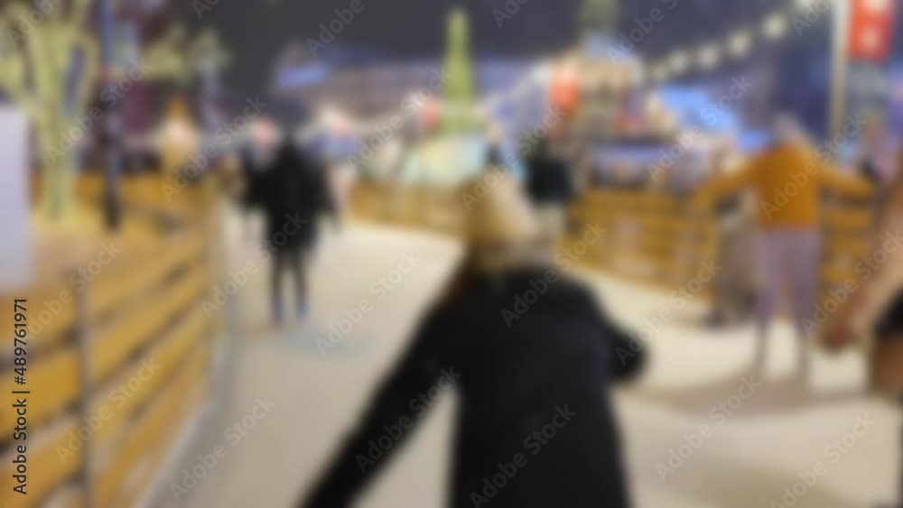 Blurred background. Many people skating on open-air ice skating rink. Snowfall on winter night. New Year Christmas. Street lights, luminous festive garland. Children family men women. City background Blurred background. Many people skating on open-air ice skating rink. Snowfall on winter night. New Year Christmas. Street lights, luminous festive garland. Children family men women. City background