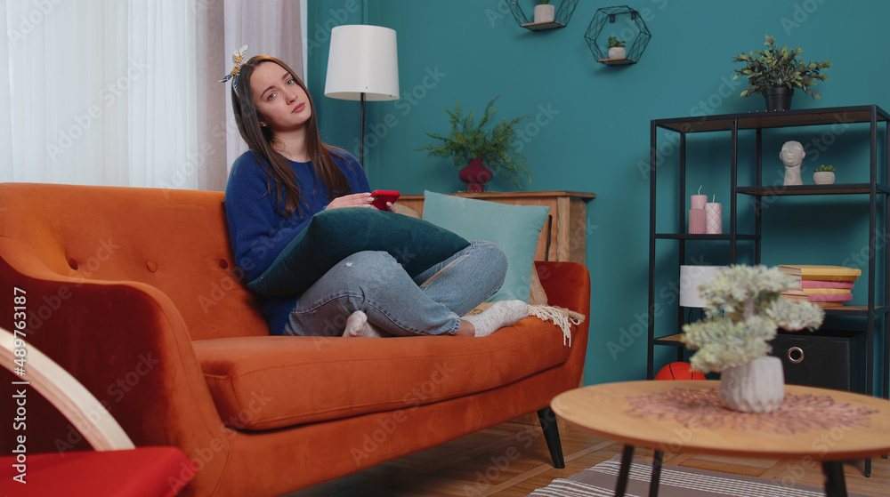 Sad teen girl in sweater use mobile cell phone typing browsing, loses becoming surprised sudden lottery results, bad fortune, loss. Young woman at home in living room on sofa. People sincere emotions Sad teen girl in sweater use mobile cell phone typing browsing, loses becoming surprised sudden lottery results, bad fortune, loss. Young woman at home in living room on sofa. People sincere emotions