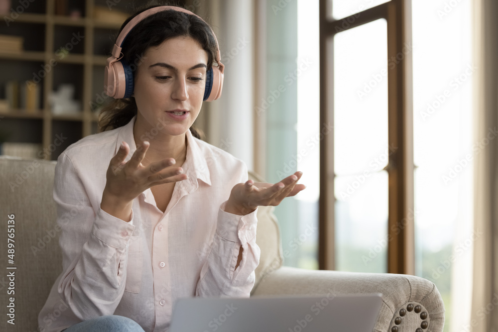 Concentrated beautiful young Hispanic woman in wireless headphones talking looking at laptop screen, involved in distant online conversation by video call application, communicating distantly at home. Concentrated beautiful young Hispanic woman in wireless headphones talking looking at laptop screen, involved in distant online conversation by video call application, communicating distantly at home.