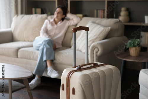 Focus on beige suitcase with blurred carefree young woman sitting on sofa on background. Joyful refreshed millennial Hispanic lady satisfied with vacation travel, relaxing alone in modern hotel room.