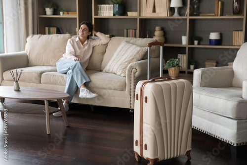 Fotografie Big beige suitcase standing on wooden floor in modern hotel room with relaxed millennial Hispanic woman sitting on comfortable couch