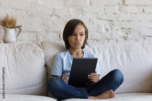 Pretty pre-adolescent 10s boy sitting on couch cross-legged holding digital tablet looking bored, spend free time alone at home. Young gen and modern tech usage, technology overuse, bad habit concept
