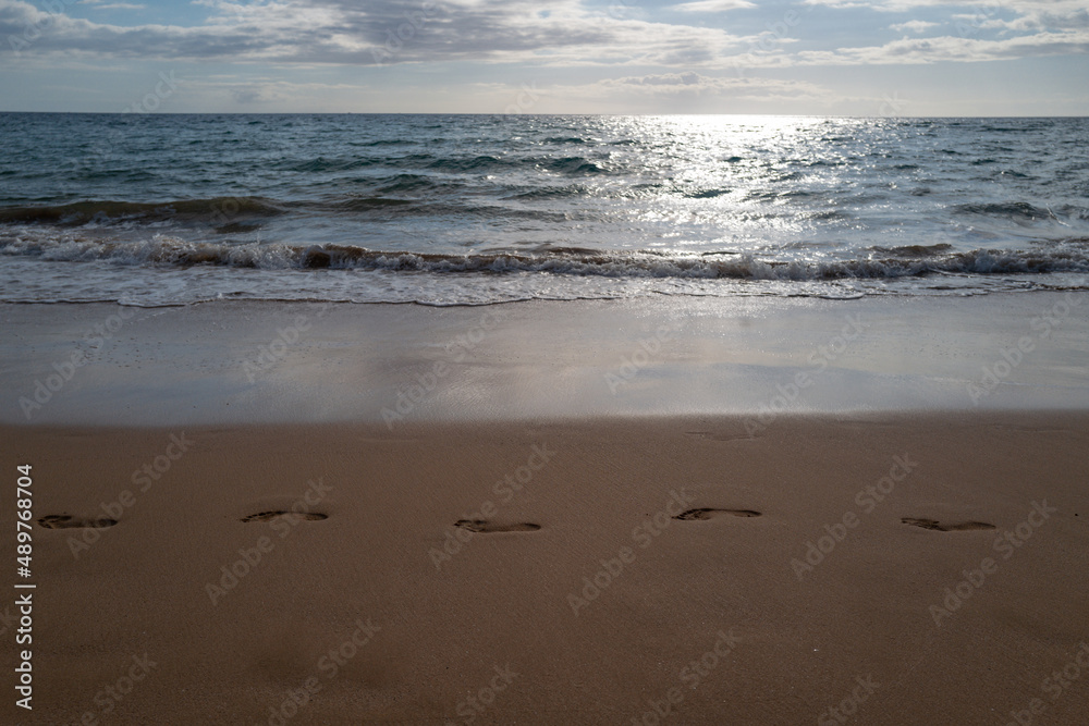 Footprints at golden sand, footsteps. Blue ocean wave on sandy beach. Beach in sunset summer time. Beach landscape. Tropical seascape, calmness, tranquil relaxing sunlight.