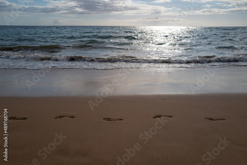 Footprints at golden sand, footsteps. Blue ocean wave on sandy beach. Beach in sunset summer time. Beach landscape. Tropical seascape, calmness, tranquil relaxing sunlight.