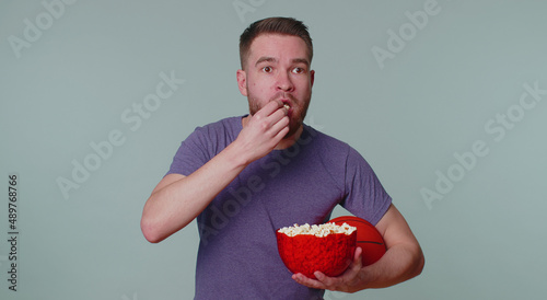 Bearded happy young man sportsman basketball fan in purple t-shirt holding and eating popcorn doing winner gesture, celebrating victory win of favourite team championship isolated on gray background