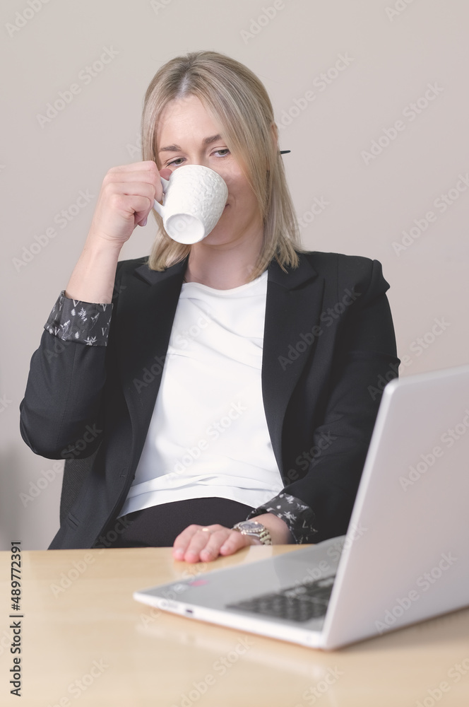 woman wearing formal suit sitting at desk with laptop and drinking coffee. having a break in the middle of a day. hard day, overworking. vertical shot. woman wearing formal suit sitting at desk with laptop and drinking coffee. having a break in the middle of a day. hard day, overworking. vertical shot.