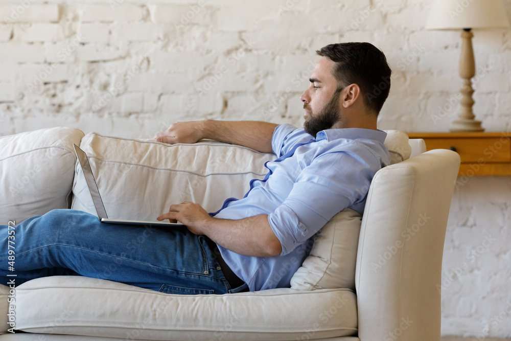 Side view serious man sit on couch with laptop, spend leisure at home on internet, buying services, ordering goods on-line, learning new software, read news in social media. Modern tech usage concept Side view serious man sit on couch with laptop, spend leisure at home on internet, buying services, ordering goods on-line, learning new software, read news in social media. Modern tech usage concept