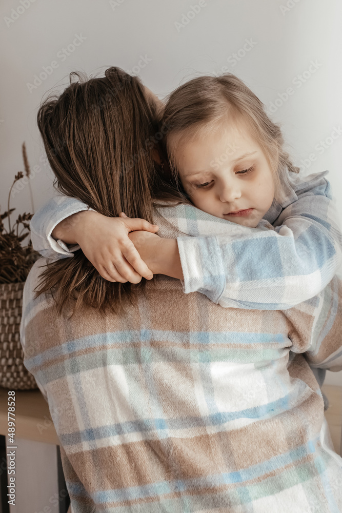 the daughter gently hugs her mother by the neck. the woman calms the girl in the sunny room.  the girl is sad