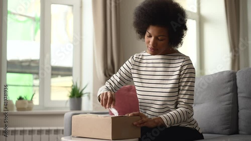 African American woman unpacking box with order and sitting on sofa in home room spbd. 4k video Closeup view of young female cuts cardboard and opens package, looks with smile and sits on couch in photo