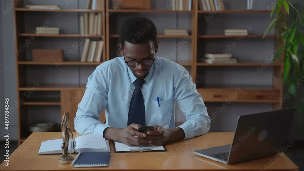 Handsome businessman using smartphone and sitting at table with laptop in company spbas. Front view of young American African man holds phone in hand and looks at screen with smile, writes message or