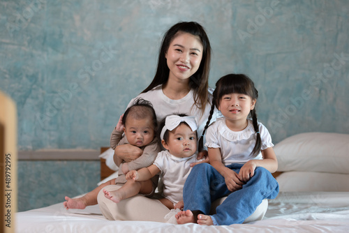Asian mom holding adorable newborn baby smile and relax in mother arm with siblings sister safety and comfortable at home.Happiness mother and her daughter sit on bed with good moment.Family concept