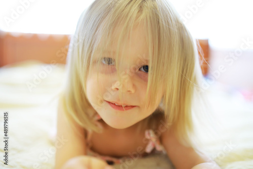 A small child with blond hair lies on the bed. Baby girl is playing on the couch.