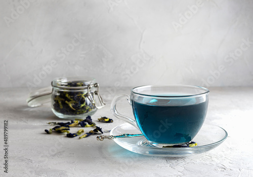Fresh tea from clitoria flowers in a transparent cup on a light table. Dried flowers in a glass jar in the background. Light background